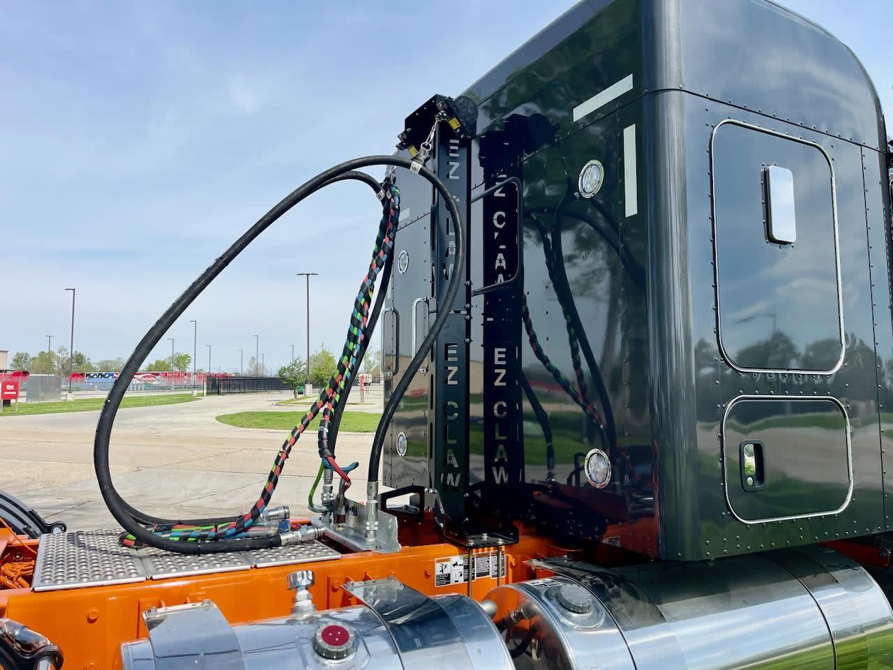 EZ Claw Center Post Mount and Hydraulic Line Saver Kit mounted to a gloss-black semi cab and trailer with an orange chassis and chrome fuel tank under clear blue sky.