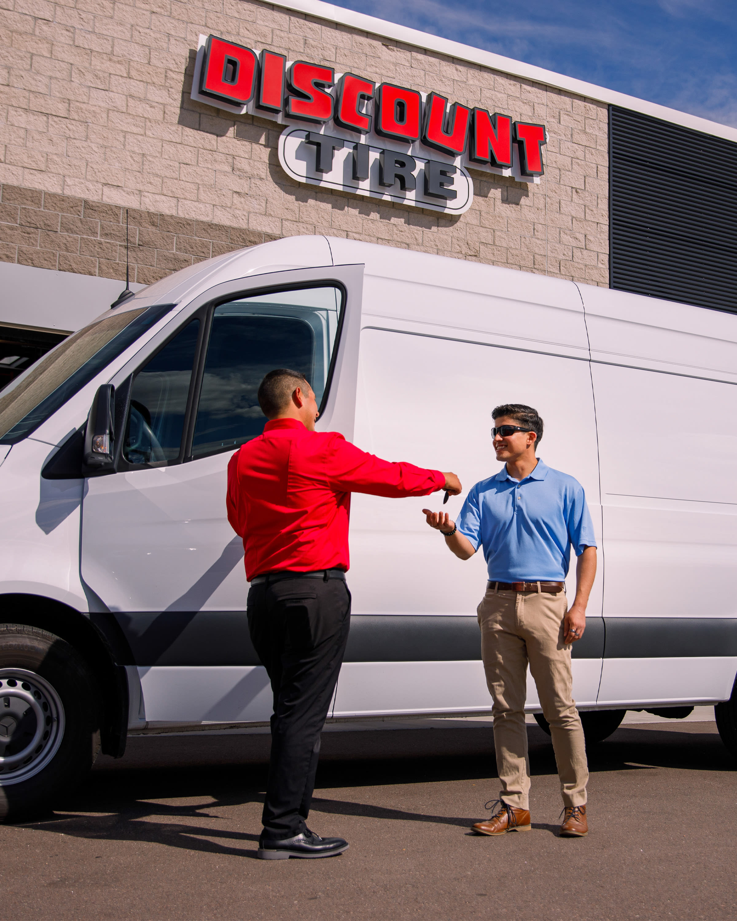 Customer and technician shake hands beside a white cargo van outside Discount Tire store, highlighting friendly service, tire sales and fleet maintenance.