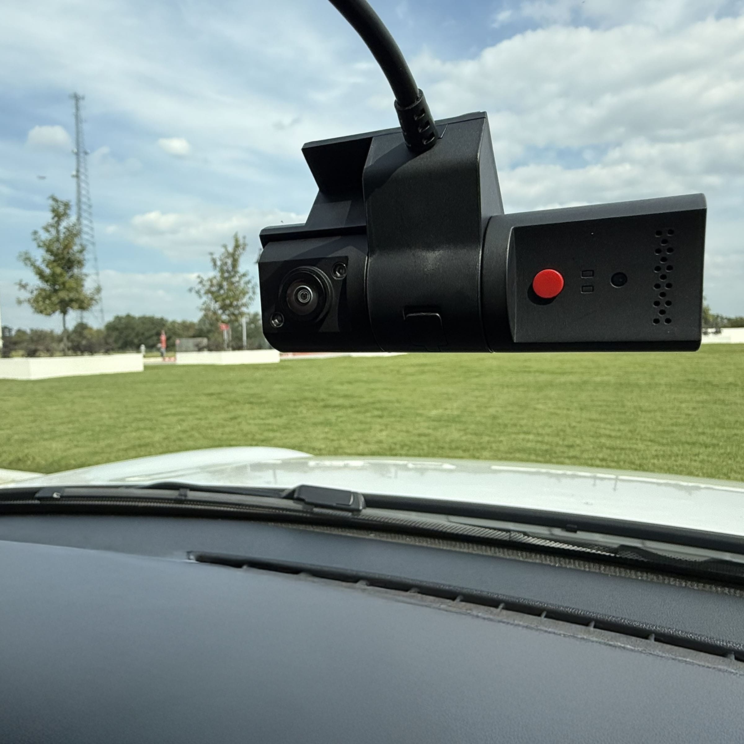 HD Fleet Dash camera mounted to upper windshield on interior vehicle. Control button including red power button is visible. A green field and trees are visible through windshield.
