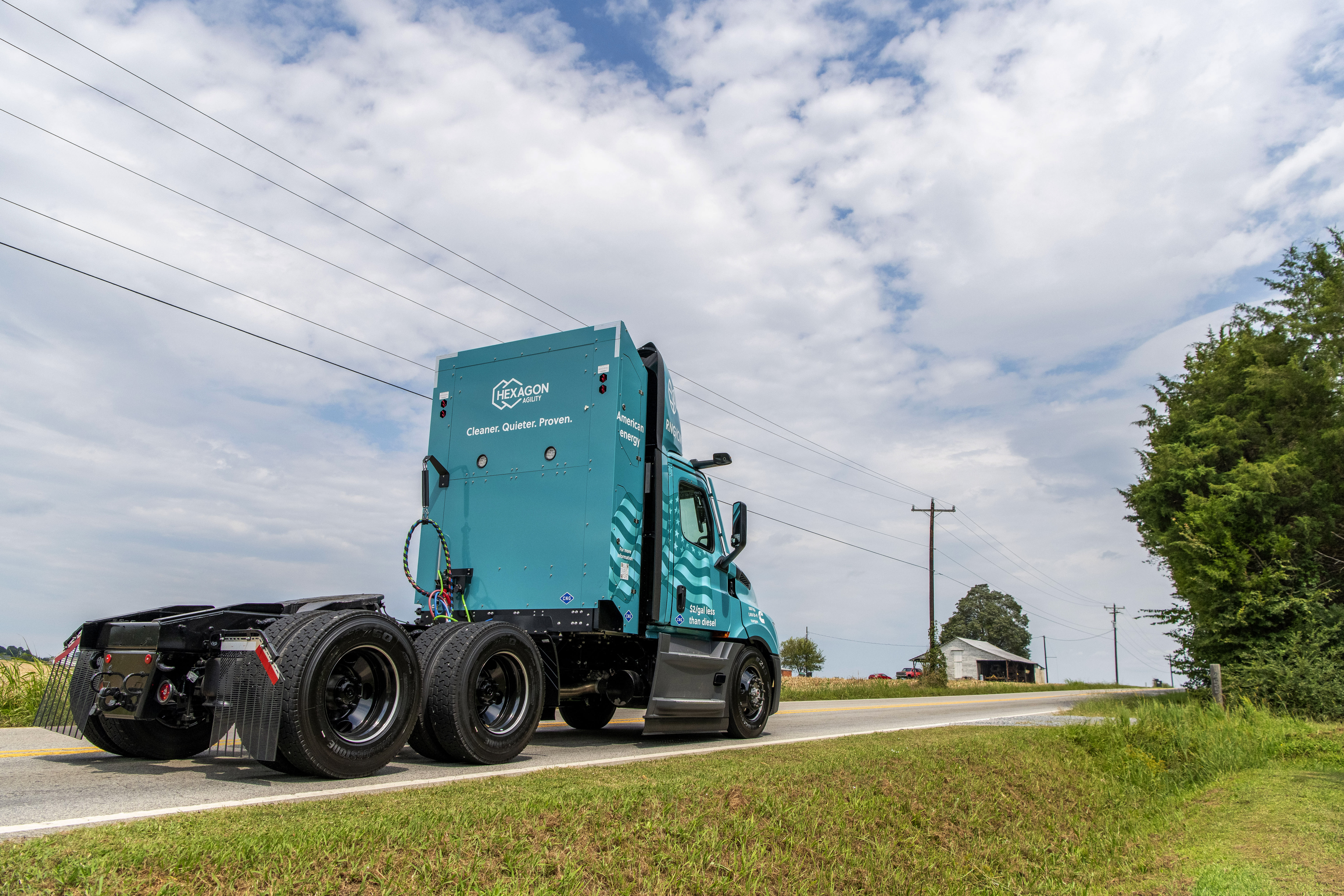 Teal semi-truck tractor cruising down a rural road under dramatic blue sky and clouds, flanked by green fields, trees and utility poles. Back side of cab has the Hexagon logo and "Cleaner. Quieter. Proven." tag line in white 