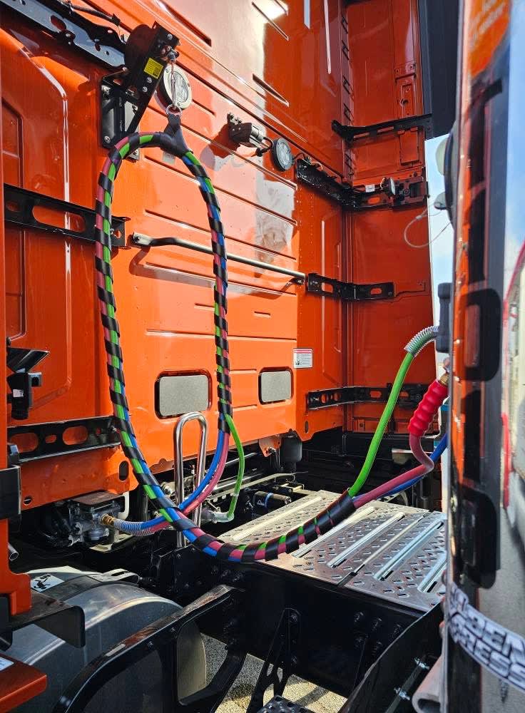 Close-up of EZ Claw Line Saver connected to orange semi-truck cabs and trailer linked by colored air lines and electrical cables, showing coupling details and polished metal steps.