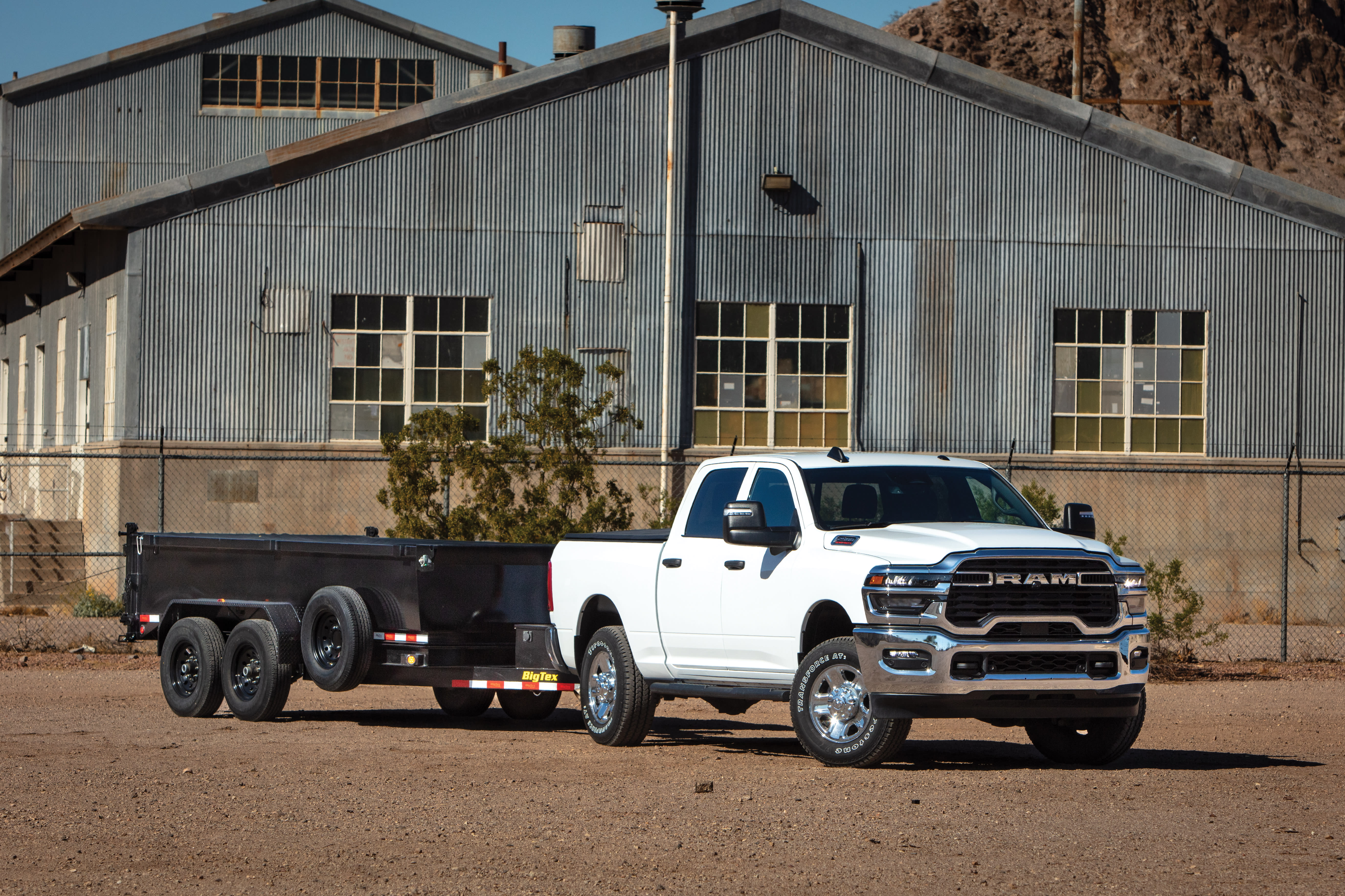 White RAM heavy-duty pickup truck towing a utility trailer in front of an industrial warehouse.