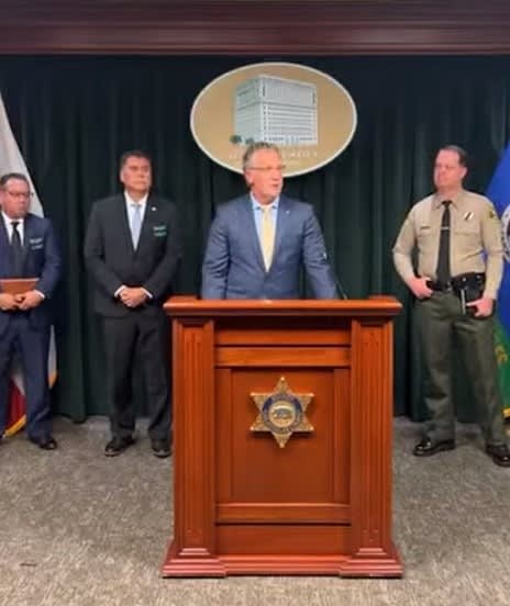 Law enforcement press conference with officials at podium with sheriff's badge