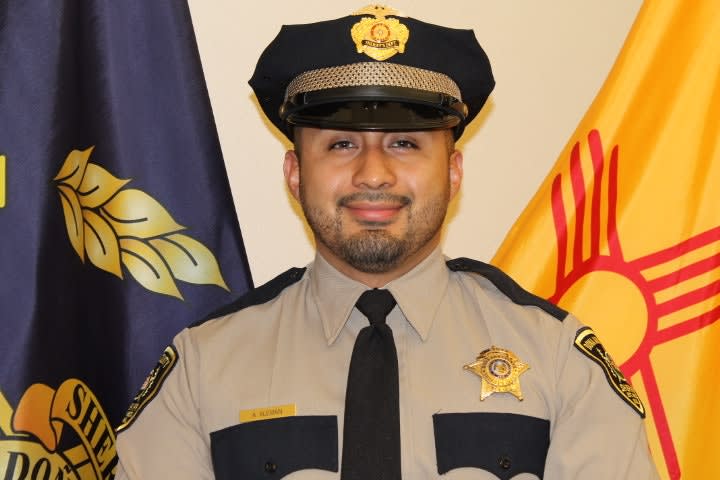 Sheriff's deputy in tan uniform and black cap, standing in front of state flags