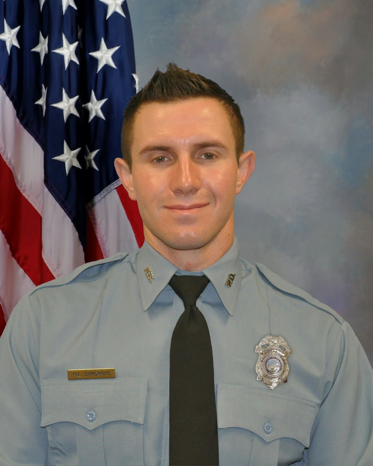 Police officer in light blue uniform standing in front of American flag