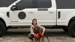 Woman kneeling with a Bloodhound in front of a white pickup truck.