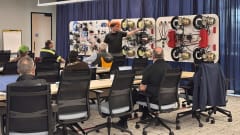 Instructor leads a brake system training class with technicians seated at tables, using a large demonstration board with air brake components and wiring.