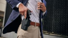 Man in a blue blazer lifting his shirt to reveal a concealed handgun in a waistband holster while standing outdoors.