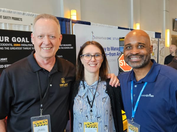 Three people smile together on the vendor expo floor, standing in front of exhibit booths and safety product displays.