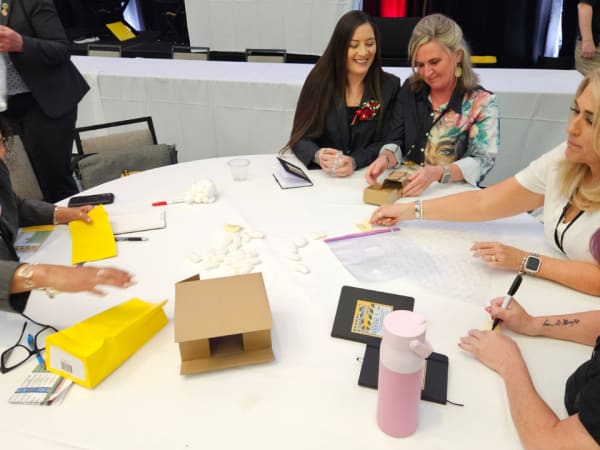 Small group of attendees participate in a hands-on workshop craft activity at a round table, using materials to assemble a paper school bus as part of a learning exercise.