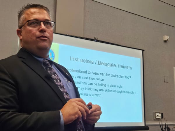 Presenter in a suit speaks during a breakout session, standing near a projection screen displaying training slides for instructors and delegate trainers.