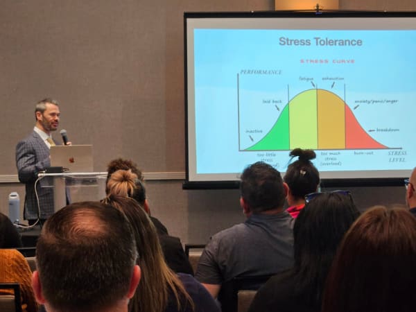 A speaker presents on stress tolerance and burnout, standing beside a projected graph illustrating performance levels and stress curves during an educational session.