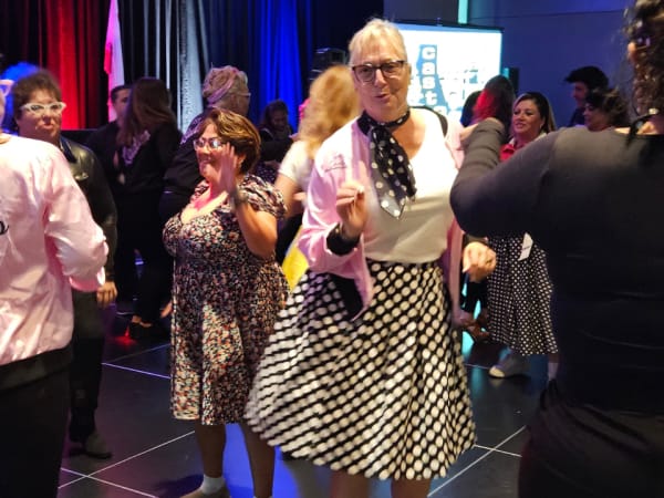 Conference attendees dressed in retro “Grease”-themed outfits dance together at the evening banquet, celebrating during the themed dinner event.