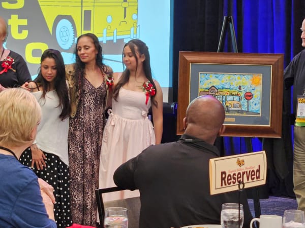 Students and presenters stand beside a framed school bus safety poster during an awards presentation recognizing contest winners.