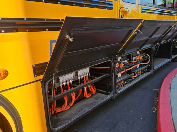 Close-up of an electric school bus with side compartments open, revealing battery and high-voltage components during an equipment demonstration.