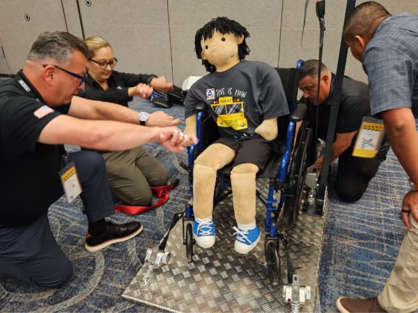 Participants practice wheelchair securement techniques using a training mannequin and tie-down equipment during a safety demonstration.