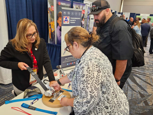 Attendees visit the AMF Bruns vendor booth in the expo hall, examining a wheelchair securement device while speaking with an exhibitor about safety equipment.