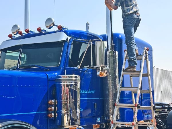 Man standing on ladder polishing exhaust stacks on a blue classic truck.