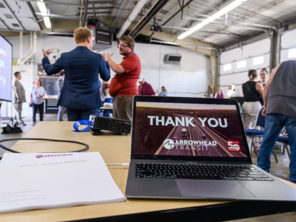 Closing slide on a laptop from the Arrowhead Transit RideSmart 2025 launch event as the crowd mingles in the background.
