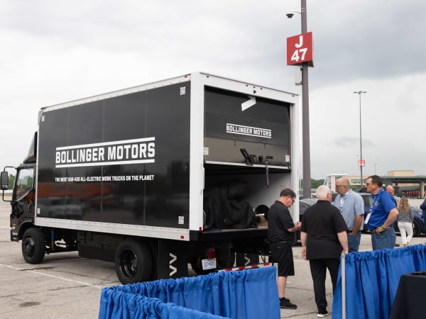 A Bollinger Motors electric truck is shown, with people chatting behind the truck.