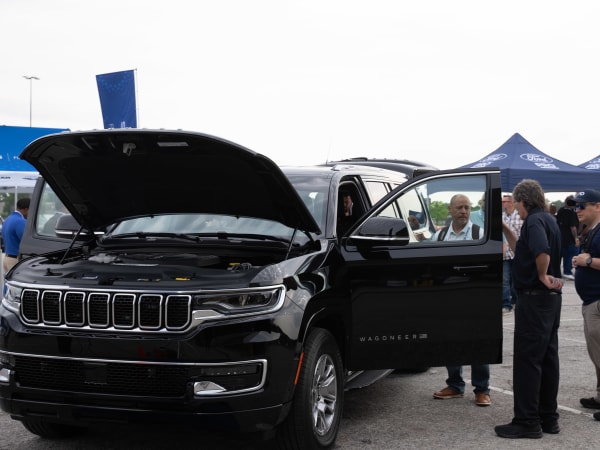The hood and driver's side door of a Jeep Wagoneer L are open as people gather around the vehicle to get a closer look at it.