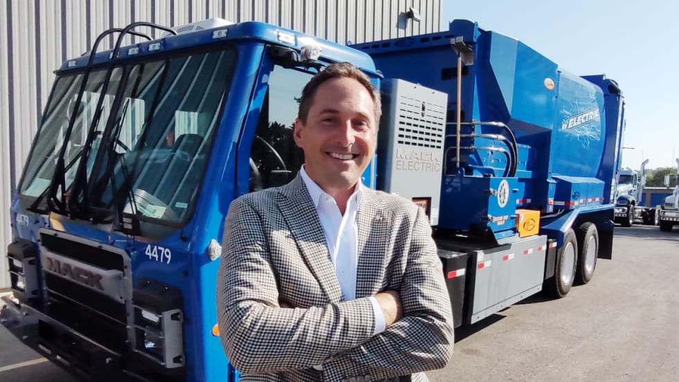 business attired man standing in front of a blue refuse truck