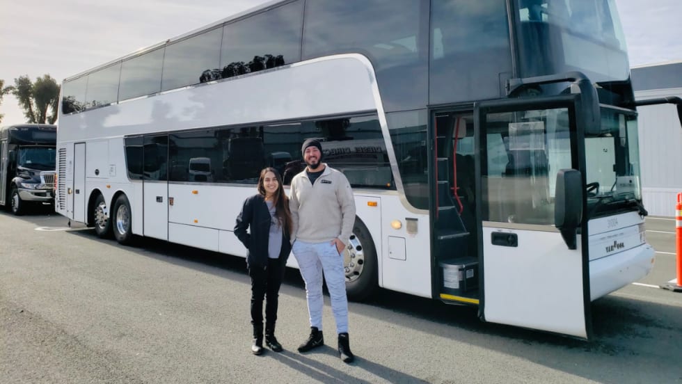 Two people standing in front of a white charter bus parked at a transportation facility.