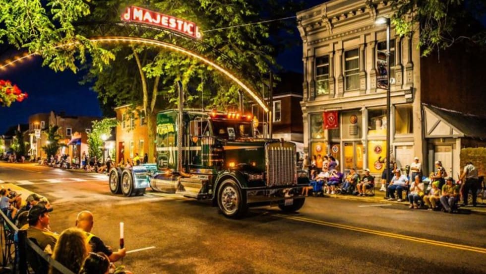 A classic Kenworth truck drives under a lit "Majestic" sign during the nighttime Kenworth Truck Parade in downtown Chillicothe, Ohio, as crowds line the street to watch the event.