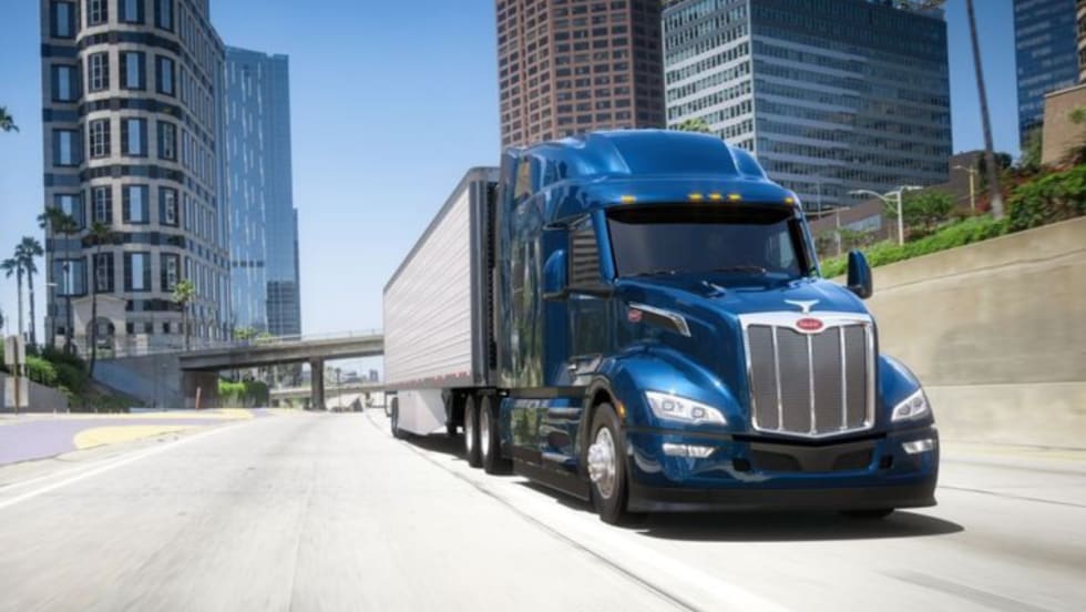 A blue Peterbilt Model 579 semi-truck driving on a city highway with tall buildings in the background.