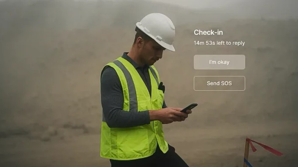 Worker in hard hat and safety vest checks phone with “I’m okay” and “Send SOS” safety check-in options on screen.
