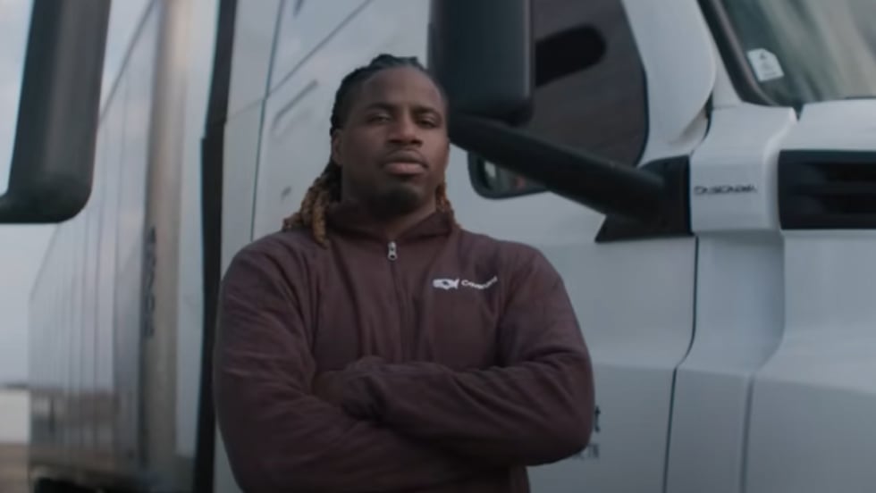 A truck driver stands with arms crossed in front of a white semi-truck, wearing a maroon jacket, featured in the ATA’s DRIVEN series highlighting professional drivers in the trucking industry.