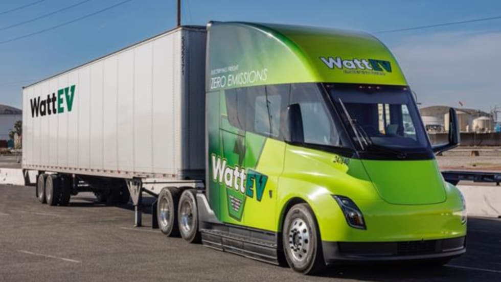 A WattEV electric semi-truck with a green cab and white trailer is parked in an industrial area under clear skies, showcasing the company's zero-emissions freight branding.