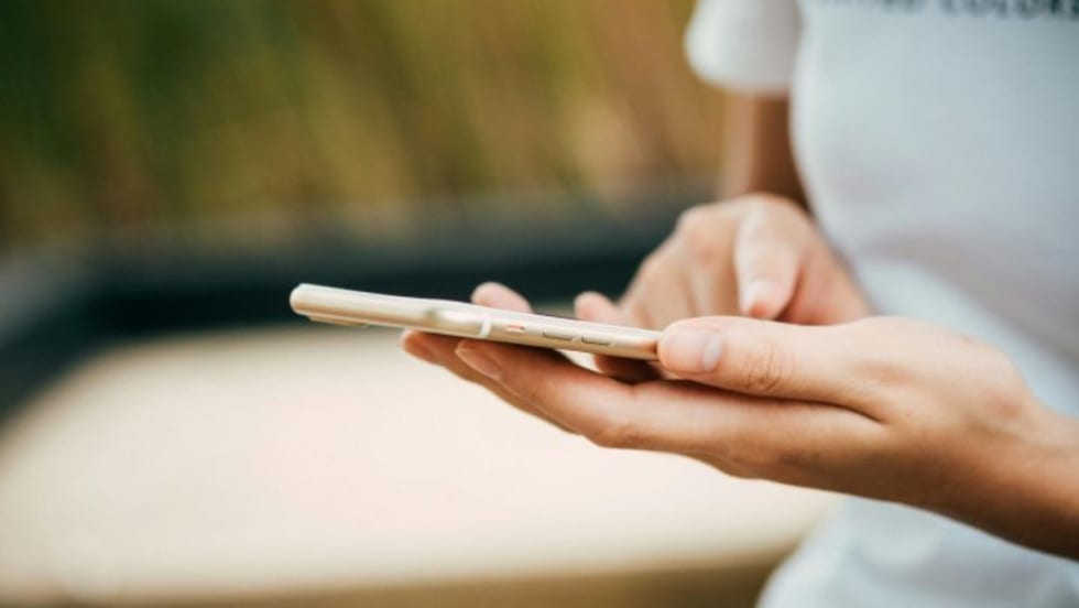Close-up of a person using a smartphone, holding it with one hand and tapping the screen with the other, wearing a white shirt with blurred outdoor background.