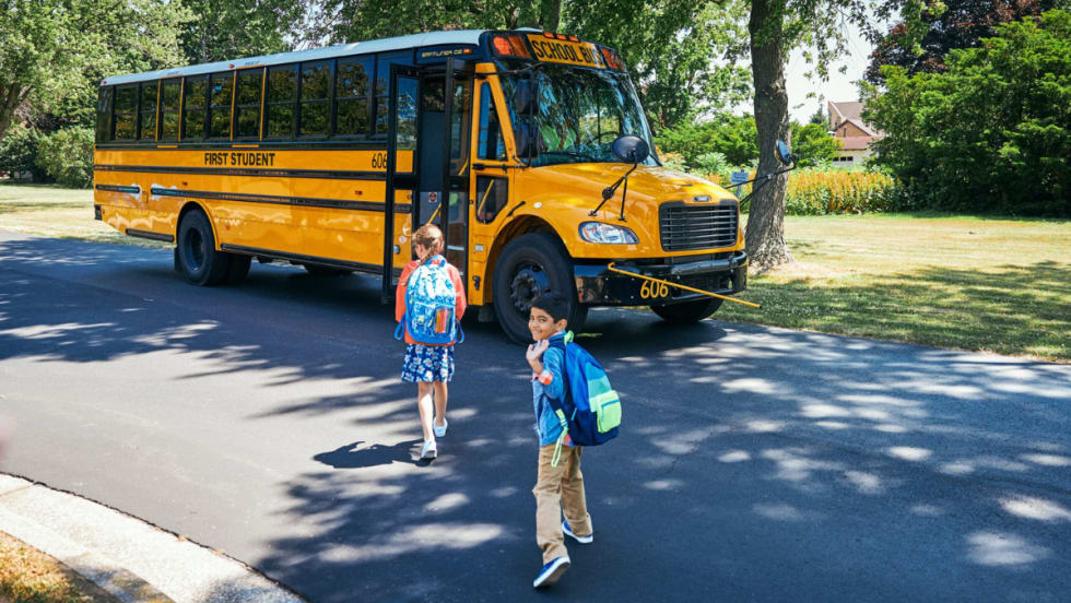 Two children with backpacks walk toward a First Student yellow school bus parked on a suburban street under the shade of trees.