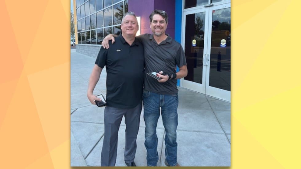 Two men stand smiling outside a building with glass doors, one with his arm around the other. Both are holding phones and dressed in casual collared shirts, one in black and one in gray.