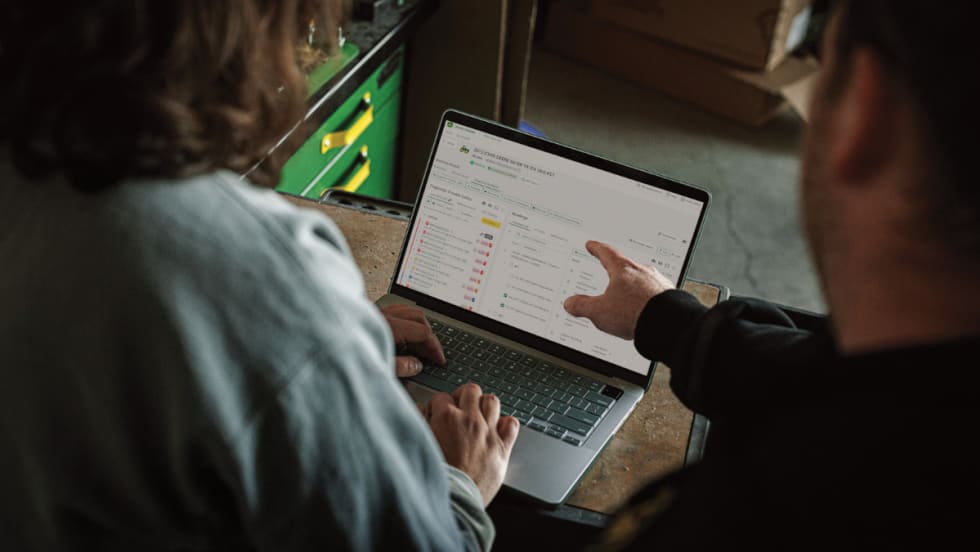 Two people review John Deere’s Operations Center PRO Service on a laptop, showing diagnostic trouble codes and repair tools for equipment maintenance.
