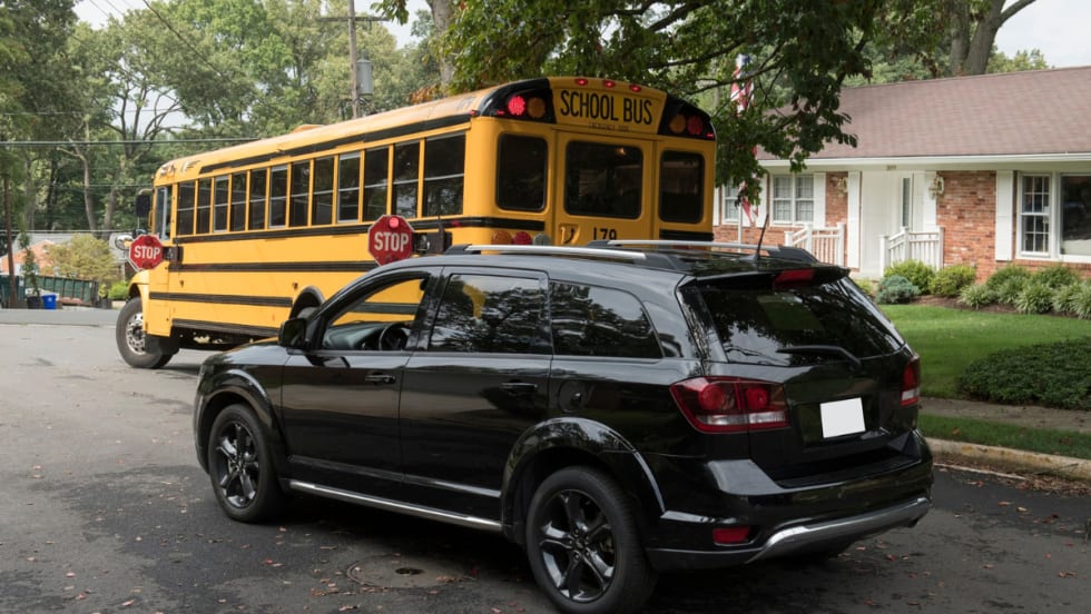 A black SUV stops behind a yellow school bus with its stop arms extended and red lights flashing in a residential neighborhood, indicating children may be boarding or exiting the bus.
