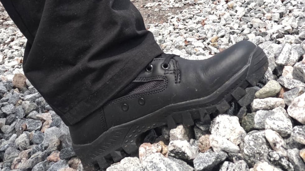 Black tactical boot with rugged sole standing on gray stones and gravel