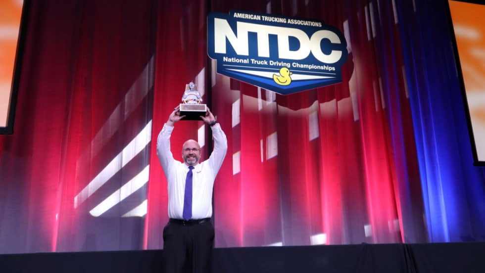 David Comings holds the NTDC Grand Champion trophy above his head on stage at the 2025 ATA National Truck Driving Championships.