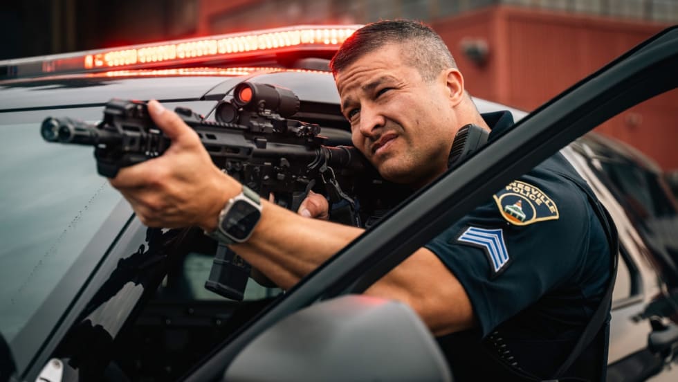 Police officer aiming tactical rifle from patrol vehicle with emergency lights