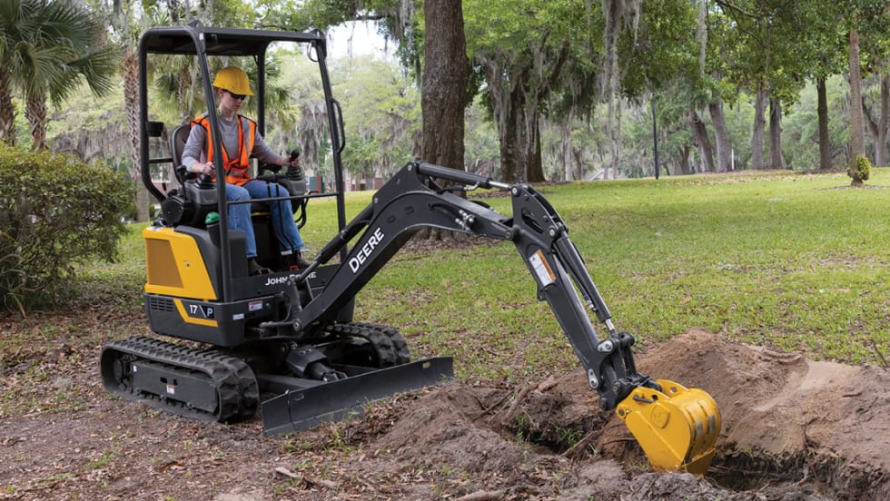 Operator using a John Deere 17 P-Tier compact excavator to dig in a grassy park area surrounded by trees.