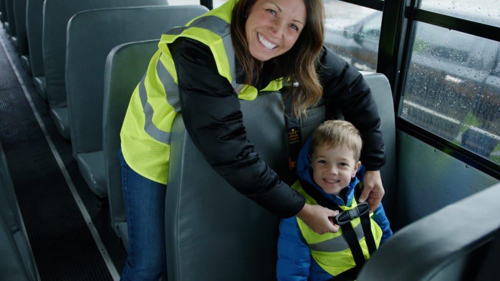 A school bus driver helps a young student buckle up while smiling inside a bus, reflecting First Student’s First Serves behavioral support program.