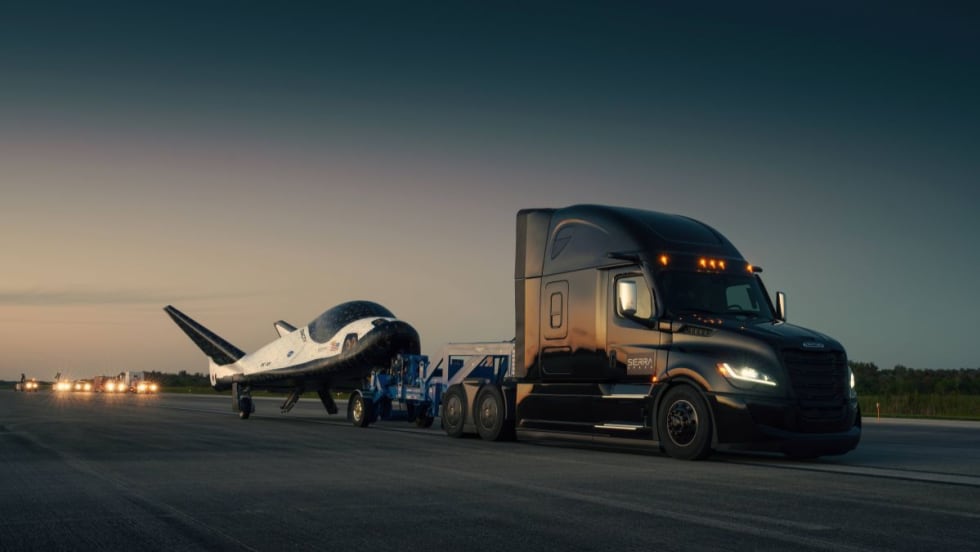 A Freightliner Cascadia semi-truck towing Sierra Space’s Dream Chaser spaceplane on a runway at dusk.