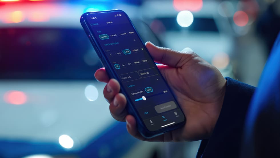 Closeup photo of man's hand holding a phone with an app displayed and out of focus police cars in the background
