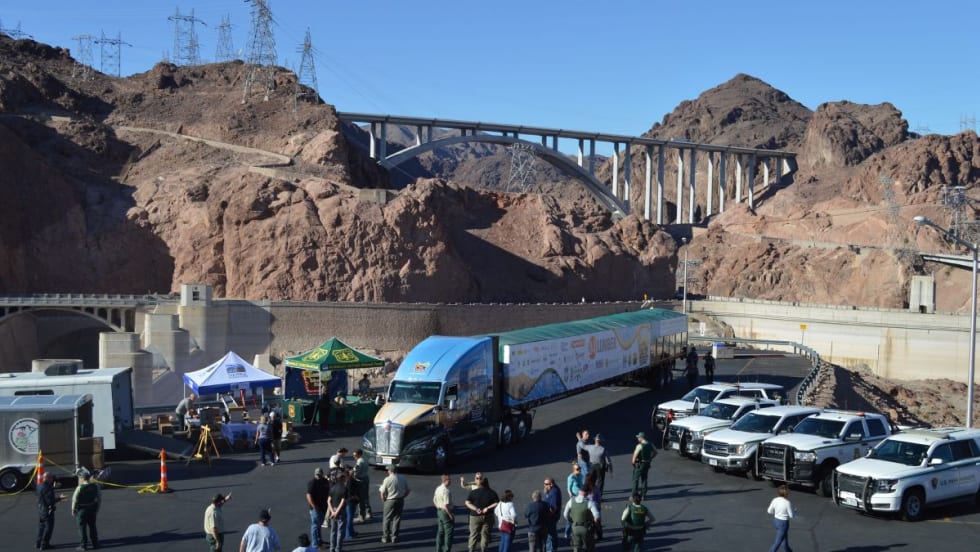 A semi-truck hauling the U.S. Capitol Christmas Tree trailer is parked near Hoover Dam, with people and law enforcement vehicles gathered nearby.