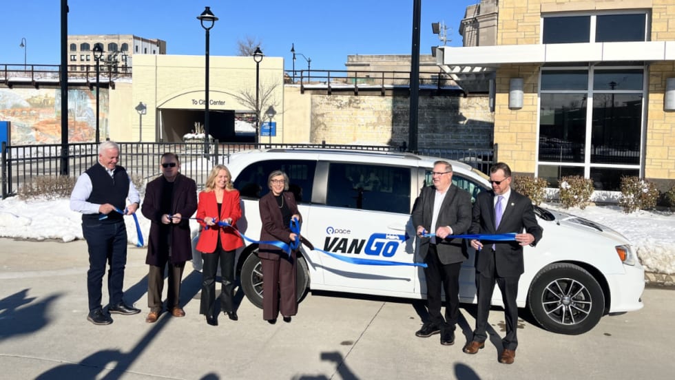 A group of officials stands in front of a white Pace VanGo minivan and holds a blue ribbon during a ribbon-cutting event outside a transit facility on a sunny winter day.