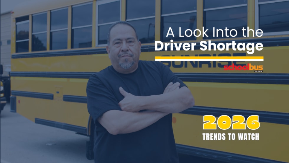 School bus driver stands with arms crossed in front of a yellow Sunrise school bus, with text reading “A Look Into the Driver Shortage” and “2026 Trends to Watch” from School Bus Fleet.