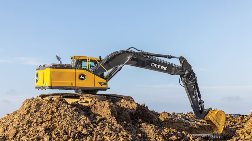 John Deere P-Tier midsize excavator positioned on a dirt mound with the boom and bucket extended at a jobsite.