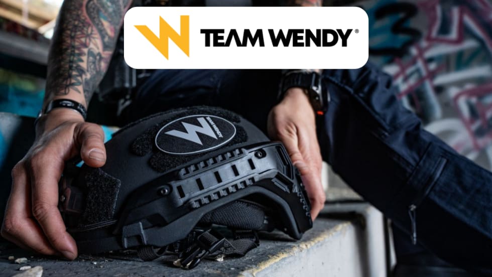 Closeup of hands reaching to pick up a ballistic helmet and a Team Wendy logo in a white box top center in the image.