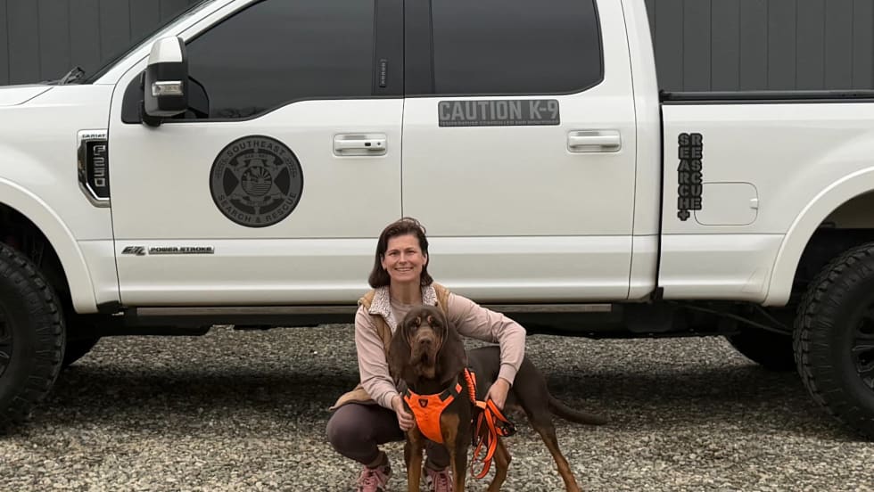 Woman kneeling with a Bloodhound in front of a white pickup truck.